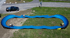 Ein blauer Bike-Park mit einer gewellten Strecke für Radfahrer. Im Hintergrund steht ein Auto auf einer graveligen Fläche.