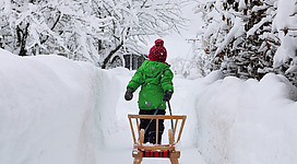 Ein Kind in einer grünen Jacke zieht einen Schlitten durch einen verschneiten Weg. Die Landschaft ist von Schnee und schneebedeckten Bäumen umgeben.