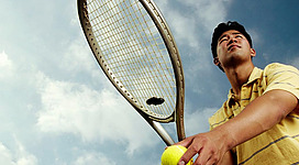Ein junger Mann hält einen Tennisschläger in der Hand und schaut nach oben. Er steht im Freien unter einem bewölkten Himmel.