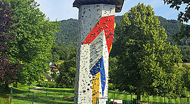 Kletterturm im Freizeitgelände Unterach am Attersee in Unterach am Attersee: Das Bild zeigt eine große freistehende Wand mit bunter Kletterwand-Struktur. Die Wand ist umgeben von grünen Bäumen und Büschen, die eine angenehme, natürliche Umgebung schaffen.