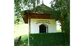 Blick auf die Kapelle hinter einer Hecke, seitlich befindet sich in der Wiese eine Holzbank