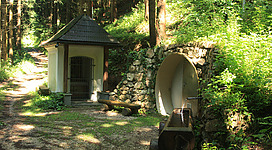 Heiliges Bründl in Auleiten in Frankenmarkt: Das Bild zeigt ein kleines, traditionelles Gebäude inmitten einer üppigen, grünen Landschaft. Das Haus hat ein spitz zulaufendes Dach und ist aus Stein gebaut. Davor befinden sich zwei große Tröge oder Brunnen, die in die natürliche Umgebung integriert sind.
