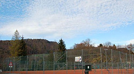 Ein Tennisplatz mit rotem Belag und umgeben von Bäumen. Der Himmel ist klar und bewölkt.
