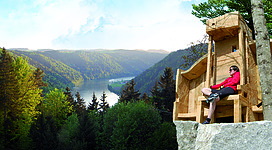 Präsidentenstuhl in Niederkappel: Eine Frau sitzt auf einer hölzernen Plattform, die sich an einem steilen Felsen befindet. Sie genießt die Aussicht auf eine bergige, bewaldete Landschaft mit einem See im Tal. Die Umgebung wirkt friedlich und idyllisch.