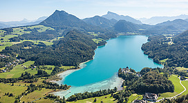 Ein malerischer See umgeben von sanften Hügeln und Bergen. Das Wasser schimmert in einem klaren Blau unter dem sonnigen Himmel.