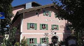 Ein charmantes, rosa Gebäude mit grünen Fensterläden und Blumenkästen. Im Vordergrund sind Tische und Stühle in einem einladenden Außenbereich.