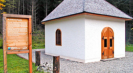 Ein kleines, weißes Gebäude mit einem Spitzdach und Holztüren steht inmitten eines Waldes. Daneben befindet sich ein informatives Schild.