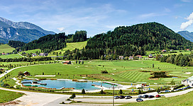 Eine beeindruckende Landschaft mit grünen Wiesen und sanften Hügeln. Im Vordergrund befindet sich ein kleiner Teich und ein Parkplatz, während im Hintergrund Berge zu sehen sind.