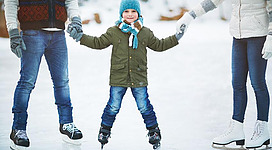 Ein Junge in skates steht auf dem Eis und hält die Hände seiner Eltern. Er trägt eine grüne Jacke, einen warmen Hut und einen Schal.