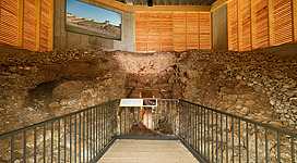 Römische Kalkbrennöfen / Calcaria in Enns: Eine beeindruckende Höhle mit mächtigen Felsen und einer klar sichtbaren Treppe, die zu einem erhöhten Bereich führt. Dort befindet sich eine große Leinwand, die offenbar einen Blick auf eine idyllische Landschaft zeigt. Die Wände der Höhle sind mit Holzpaneelen verkleidet, die ihr ein gemütliches Ambiente verleihen.