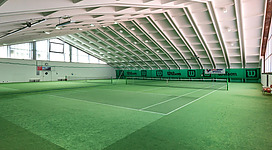 Eine Tennis-Halle mit grünem Teppichboden und einem großen, hellen Dach. Im Hintergrund sind Tennisschläger und Werbeplakate sichtbar.