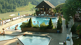 Freibad Sankt Nikola in St. Nikola an der Donau: Ein Resort-Komplex mit einem großen Außenpool, der von Liegestühlen und Sonnenschirmen umgeben ist. Das zweistöckige Hauptgebäude hat ein Satteldach und eine offene Veranda. Die Umgebung ist geprägt von üppigem Grün mit Bäumen und Sträuchern, die eine malerische Kulisse bilden.