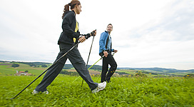 Fitness Parcour in Lohnsburg am Kobernaußerwald: Zwei Personen laufen mit Wanderstöcken über eine grüne Wiese. Sie tragen Sportbekleidung und scheinen sich auf einer Wanderung oder Exkursion zu befinden. Die Landschaft im Hintergrund ist hügelig und grün.
