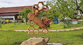 Bienenlehrpfad in Franking in Franking: Eine große, dreidimensionale Skulptur aus Holz, die verschiedene Bienen und Waben in einem geometrischen Muster darstellt. Die Skulptur ist auf einem Felsen in einer grünen Wiese platziert, umgeben von Bäumen. Es scheint sich um eine künstlerische Darstellung von Bienen und Honigwaben zu handeln.