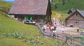 Eine malerische Landschaft mit traditionellen Holzhäusern. Menschen versammeln sich im Freien, umgeben von grünen Wiesen und Hügeln.