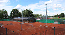 Tennisplatz in Andorf: Eine Reihe von Tennisplätzen mit rötlichen Sandböden sind von Gittern umgeben. In der Ferne sind Bäume und ein Gebäude zu sehen, wahrscheinlich eine Umkleidekabine oder ein Clubhaus. Insgesamt ein sportliches und gut gepflegtes Outdoor-Tennisgelände.