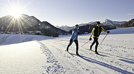 Zwei Personen laufen auf Langlaufskiern durch eine schneebedeckte Landschaft. Im Hintergrund sind Berge und ein strahlender Sonnenaufgang zu sehen.