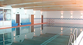 Ein ruhiger Innenpool mit glattem Wasser und Holzdecke. Die Wände sind in hellen Farben gehalten und es gibt blaue Sitzgelegenheiten.