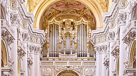 Brucknerorgel in St. Florian: Eine prächtige Kirche mit reich verzierter Fassade und Orgelprospekt. Die Architektur ist im prunkvollen Barockstil gehalten, mit hohen Bögen, Säulen und einer üppigen Ornamentik. Der Raum ist hell erleuchtet und strahlt eine feierliche Atmosphäre aus.