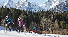 Eine Familie zieht Schlitten durch den Schnee in den Bergen. Im Hintergrund sind hohe, schneebedeckte Gipfel und Bäume zu sehen.