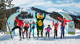 Eine Gruppe von Skifahrern steht auf einer verschneiten Piste und posiert fröhlich. Ein Maskottchen in einem grünen Kostüm hebt seine Arme in die Luft.