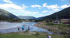 Ein ruhiger Fluss umgeben von grünen Hügeln und Bäumen. Menschen genießen das Wasser und die Natur.