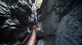 Eine enge Schlucht mit hohen Felswänden und einem Holzsteg. Einige Personen gehen den Weg entlang, während das Licht von oben strahlt.