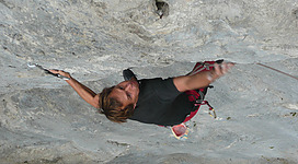 Ein Sportkletterer wird an einer steilen Felswand gesehen. Er greift nach einem Steinfelsen, während er sich vertikal bewegt.