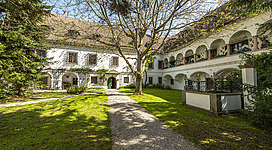 Schloss Weyer in Gmunden: Das Bild zeigt ein historisches Gebäude mit charakteristischer Architektur. Es ist von einem gepflegten Garten mit Rasenfläche und Bäumen umgeben, was dem Ensemble einen landschaftlichen Charme verleiht.
