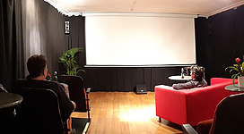 Ein gemütlicher Raum mit einer Leinwand und Zuschauerplätzen. Die Wände sind schwarz und es gibt einige Pflanzen in der Ecke.