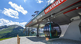 Eine moderne Seilbahnstation in den Bergen mit kabinen. Im Hintergrund sind grüne Hügel und blauer Himmel zu sehen.