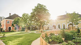 Stiegl-Gut Wildshut (Schaubetrieb - Brauerei ) in St. Pantaleon: Ein historisches Gebäude umgeben von üppiger Vegetation und einem gepflegten Garten. Eine Terrasse mit Möbeln lädt zum Verweilen ein. Die Sonne scheint durch die Blätter der Bäume und wirft ein warmes Licht auf die Szenerie.