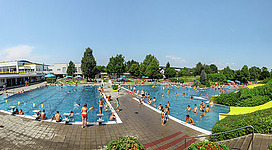 Vitalwelt Perg in Perg: Ein großer und lebendiger Außenpools mit vielen Menschen, die darin schwimmen und sich sonnen. In der Umgebung befinden sich Bäume und Grünanlagen.