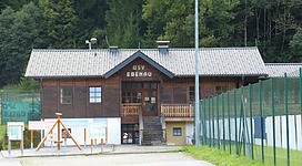 Ein traditionelles Holzgebäude mit einem Zeltplatz in der Nähe. Umgeben von Bäumen und einem Sportplatz.
