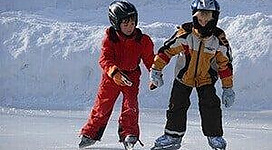 Zwei Kinder stehen auf einer Eisbahn und halten sich an den Händen. Sie tragen Helme und warme Kleidung, während sie das Eislaufen lernen.