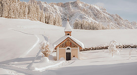Eine kleine Kirche in einer schneebedeckten Landschaft. Im Hintergrund sind majestätische Berge und ein klarer Himmel sichtbar.