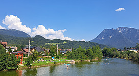 Ein Fluss mit klarer Wasseroberfläche, in dem mehrere Menschen schwimmen und paddeln. Am Ufer liegen Personen auf einer grünen Wiese, einige mit Sonnenschirmen. Dahinter Häuser, Wälder und hohe Berge unter blauem Himmel mit weißen Wolken. Sommerliche, idyllische Landschaft.