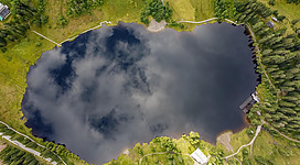 Ein ruhiger Teich umgeben von grünen Wiesen und Bäumen. Die Wolken spiegeln sich sanft auf der Wasseroberfläche.