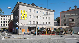 Ein modernes Gebäude mit auffälliger Beschriftung und Außenbereich für Gäste. Die Straße ist belebt und zeigt eine Mischung aus modernen und historischen Architektur.