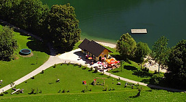 Ein kleines gelbes Haus liegt am Ufer eines klaren Sees, umgeben von Bäumen. Rund um das Haus stehen Liegen und Sonnenschirme auf einer grünen Wiese.