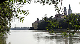 Naturschutzgebiet "Vornbacher Enge" in Wernstein am Inn: Eine malerische, mittelalterliche Abtei mit Kirchtürmen, umgeben von grünen Bäumen und einem ruhigen See, der die Spiegelung des Gebäudes widerspiegelt. Die Szene vermittelt eine friedliche, romantische Atmosphäre inmitten der Natur.