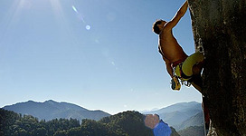 Ein Kletterer, der an einer Felswand in den Bergen klettert. Im Hintergrund sind Wälder und Hügel unter einem strahlend blauen Himmel zu sehen.