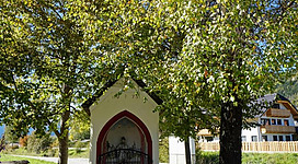 Eine kleine Kapelle umgeben von großen Bäumen. Im Hintergrund sind mehrere Gebäude sichtbar.