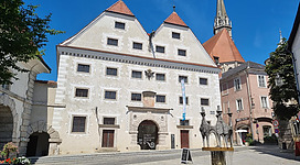 Stadtmuseum Steyr in Steyr: Das Bild zeigt ein historisches Gebäude mit markanten Türmen und aufwendig verzierter Fassade. Der Platz davor ist mit einem Brunnen und Bäumen gestaltet. Das Gebäude scheint ein Museum oder öffentliches Gebäude zu sein.