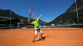 Ein Junge spielt Tennis auf einem Sandplatz. Im Hintergrund sind grüne Berge und ein strahlend blauer Himmel zu sehen.