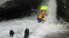 Eine Person mit einem grünen Helm und einer Schwimmweste sitzt im sprudelnden Wasser. Es sieht so aus, als würde sie von einem Wasserfall umgeben sein und hat viel Spaß.
