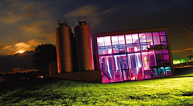 Ein modernes Gebäude mit großen Fenstern, das in bunten Neonfarben beleuchtet ist. Im Hintergrund stehen zwei silberne Silos auf einer grünen Wiese.