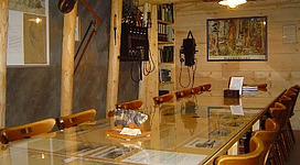 Bergbaumuseum in St. Pantaleon: Ein geräumiger Innenraum mit Holzvertäfelung und -möbeln, dekoriert mit allerlei Exponaten und Utensilien. Ein großer Esstisch steht im Mittelpunkt, umgeben von Stühlen. An den Wänden hängen Gemälde und Fotos. Alles wirkt gemütlich und einladend.