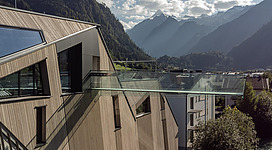 Ein modernes Gebäude mit einer Glasveranda, das in die Berge blickt. Die Umgebung ist von beeindruckenden Berglandschaften und blauem Himmel geprägt.