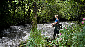 Angeln im Rannatal in Oberkappel: Ein Mann steht an einem reißenden Fluss, umgeben von üppigem, grünem Wald. Er hält eine Angelrute und scheint sich auf das Angeln zu konzentrieren. Die Umgebung wirkt idyllisch und ruhig, mit Blumen am Flussufer.